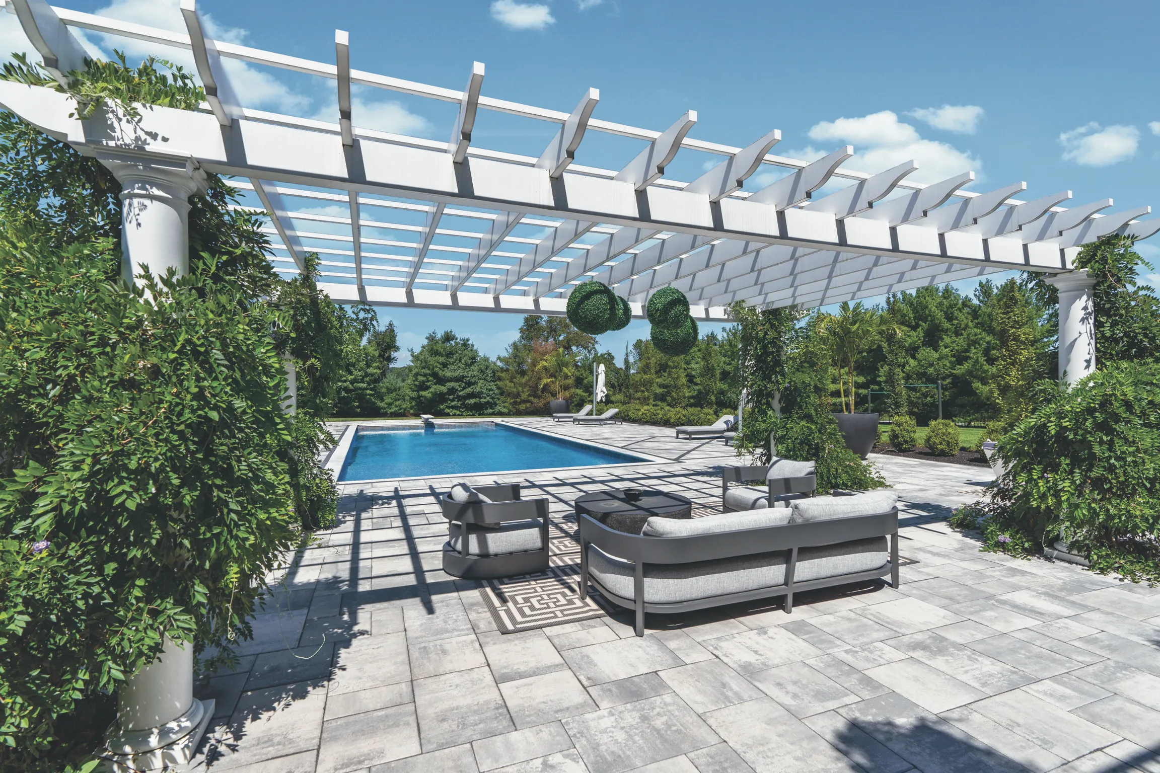 Modern poolside lounge area with a white pergola, stylish outdoor furniture, and lush landscaping under a clear blue sky.