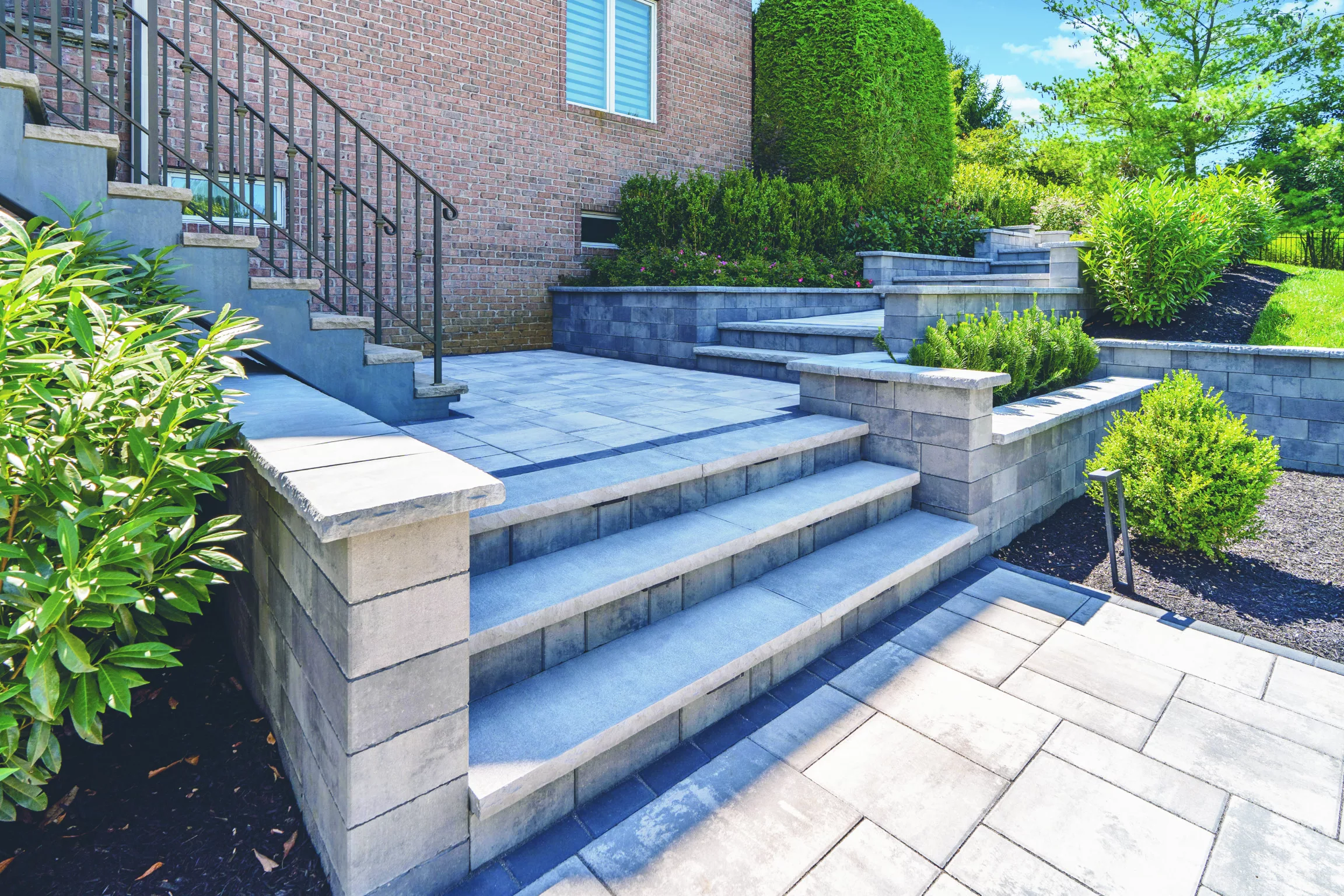 Elegant gray stone steps and patio area with integrated planters, surrounded by lush landscaping next to a brick house.