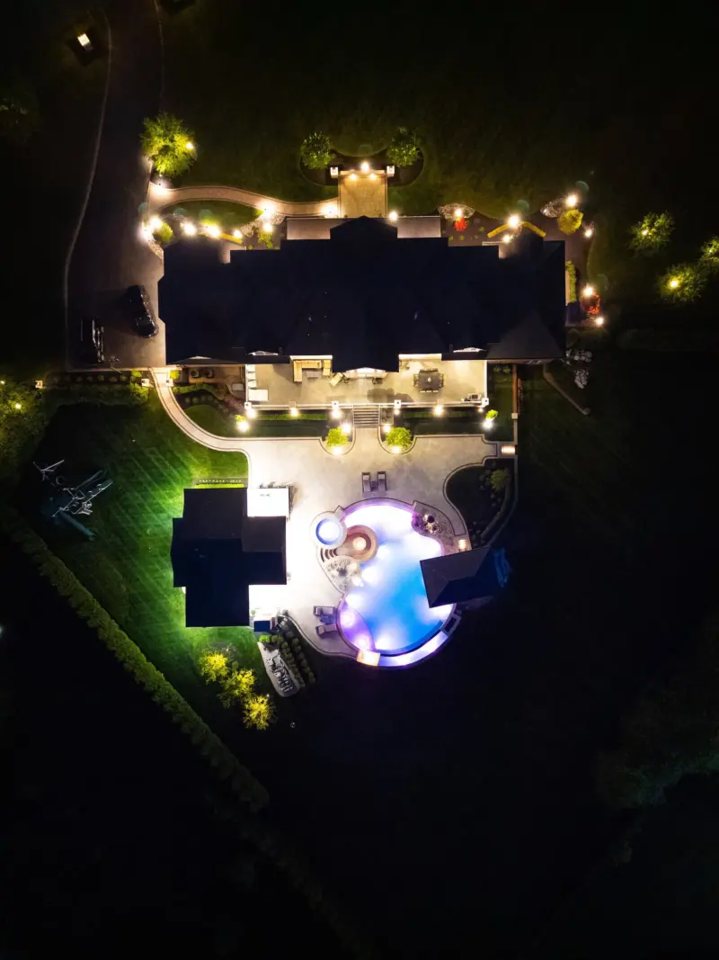 Aerial night view of freeform pool, raised spa, travertine patio, gas fire pit, and landscape lighting on a Colts Neck NJ estate