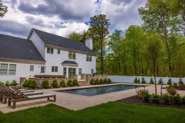 A full backyard transformation featuring a raised patio with stone retaining wall and pillars, paver pool patio with coping, paver steps with integrated LED lighting, a paver walkway with path lights, Japanese maple specimen plantings, arborvitae privacy screening, and sod installation.