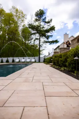 A full backyard transformation featuring a raised patio with stone retaining wall and pillars, paver pool patio with coping, paver steps with integrated LED lighting, a paver walkway with path lights, Japanese maple specimen plantings, arborvitae privacy screening, and sod installation.