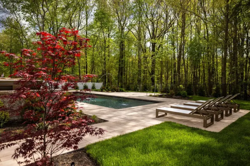 Paver pool patio with Japanese maple, lounge chairs, planting beds, and pool with fountain jets on wooded lot in Old Bridge NJ