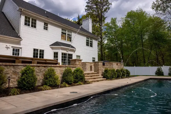 A full backyard transformation featuring a raised patio with stone retaining wall and pillars, paver pool patio with coping, paver steps with integrated LED lighting, a paver walkway with path lights, Japanese maple specimen plantings, arborvitae privacy screening, and sod installation.