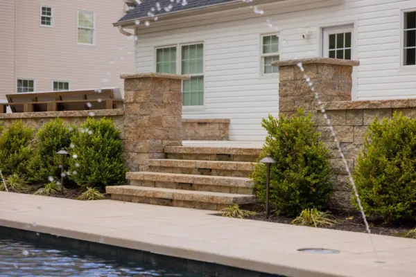 A full backyard transformation featuring a raised patio with stone retaining wall and pillars, paver pool patio with coping, paver steps with integrated LED lighting, a paver walkway with path lights, Japanese maple specimen plantings, arborvitae privacy screening, and sod installation.