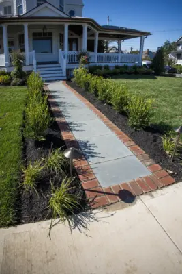 Scenic View Landscaping transformed a Spring Lake property with a complete front and backyard renovation, featuring beach-inspired landscaping, custom lighting, bluestone patio, brick driveway, outdoor kitchen, new sod, drainage, and concrete walkways.