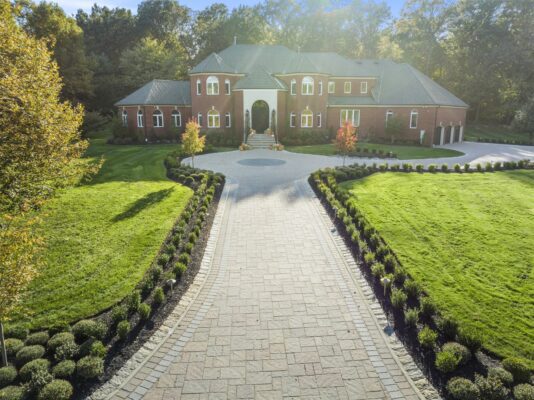 Straight-on view of a custom brick paver driveway entrance with border detail and landscaped beds leading to a home in Colts Neck NJ