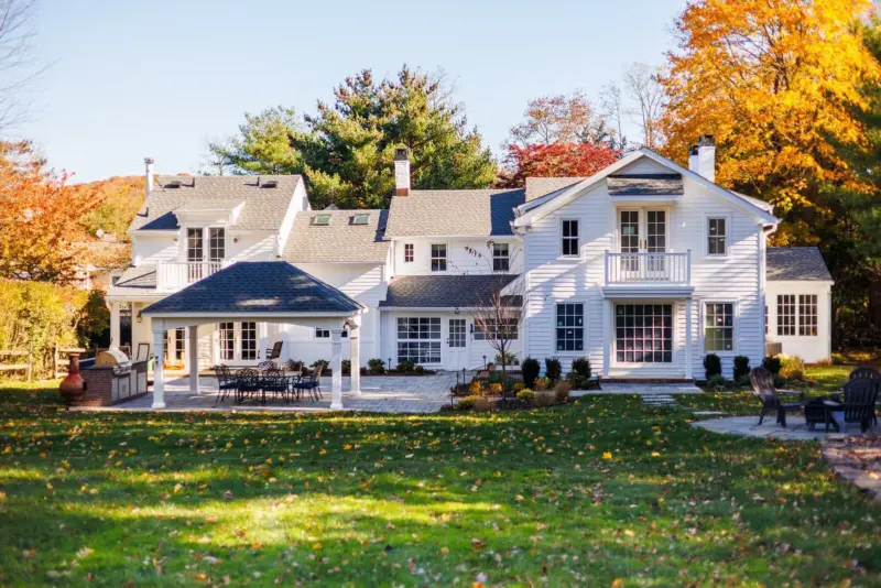Rear view of a white colonial home in Rumson, NJ with a covered patio, paver patio, walkway, and fire pit area