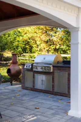 A covered paver patio with built-in outdoor kitchen, stone steps, natural stone walkway, and a dedicated fire pit area tied together with plantings and landscape lighting.