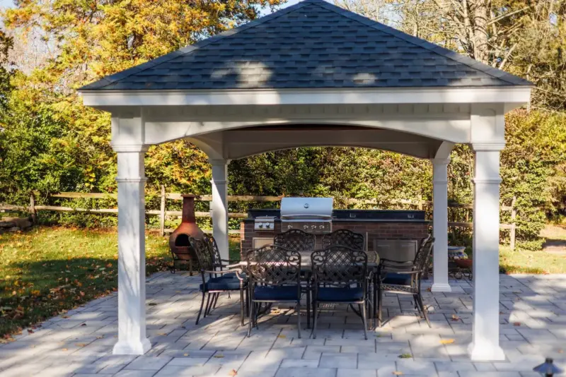 White covered patio pavilion with a shingled roof, dining set, and built-in outdoor kitchen on a paver patio in Rumson, NJ