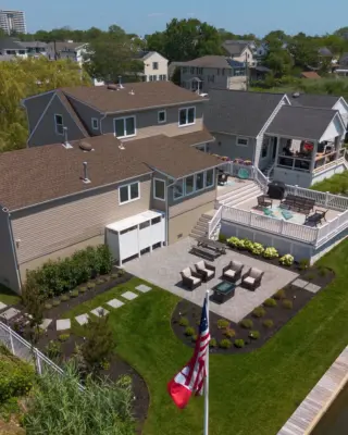 A herringbone paver patio and fire table lounge below the deck, with a bluestone stepping stone path, curved planting beds, new sod, and lighting leading to the water.