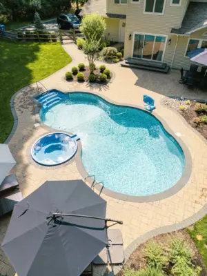 A paver pool patio wrapping a freeform pool and spa, a separate circular fire pit patio with built-in stone fire pit, a curved paver walkway, and refreshed planting beds on a wooded lot.