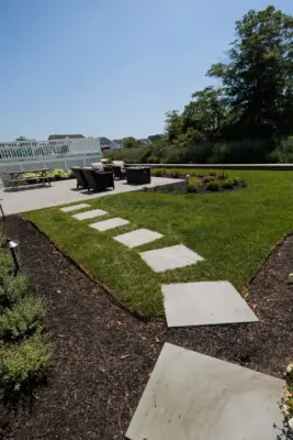 A herringbone paver patio and fire table lounge below the deck, with a bluestone stepping stone path, curved planting beds, new sod, and lighting leading to the water.