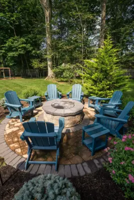 A paver pool patio wrapping a freeform pool and spa, a separate circular fire pit patio with built-in stone fire pit, a curved paver walkway, and refreshed planting beds on a wooded lot.