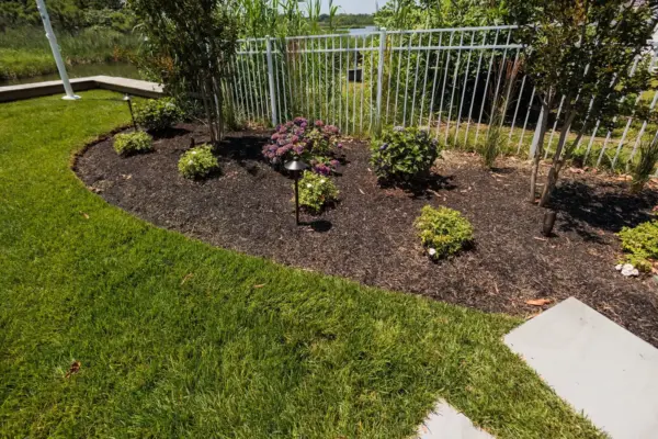 A herringbone paver patio and fire table lounge below the deck, with a bluestone stepping stone path, curved planting beds, new sod, and lighting leading to the water.