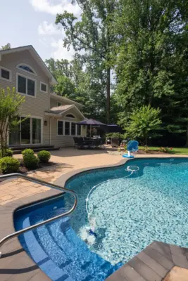 A paver pool patio wrapping a freeform pool and spa, a separate circular fire pit patio with built-in stone fire pit, a curved paver walkway, and refreshed planting beds on a wooded lot.