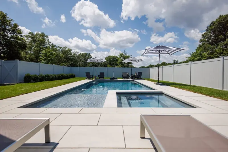 Ground-level view of modern rectangular pool and spa with large-format paver patio, lounge chairs, sod lawn and vinyl fence in Oceanport, NJ