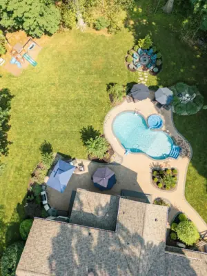 A paver pool patio wrapping a freeform pool and spa, a separate circular fire pit patio with built-in stone fire pit, a curved paver walkway, and refreshed planting beds on a wooded lot.