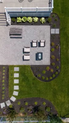 A herringbone paver patio and fire table lounge below the deck, with a bluestone stepping stone path, curved planting beds, new sod, and lighting leading to the water.