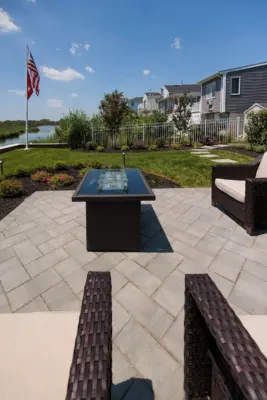A herringbone paver patio and fire table lounge below the deck, with a bluestone stepping stone path, curved planting beds, new sod, and lighting leading to the water.