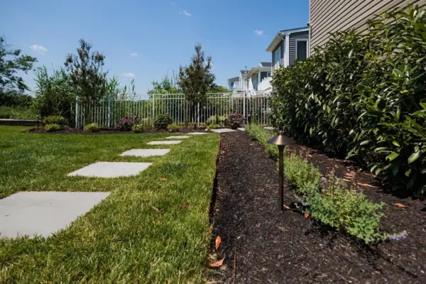 A herringbone paver patio and fire table lounge below the deck, with a bluestone stepping stone path, curved planting beds, new sod, and lighting leading to the water.