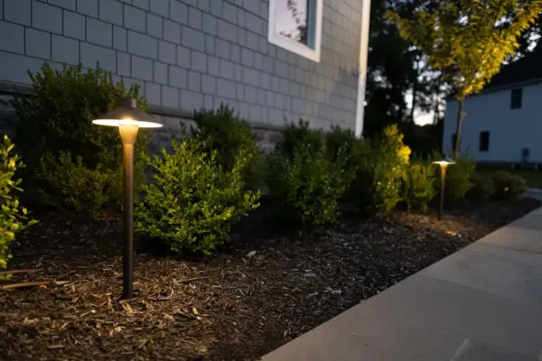 A complete outdoor transformation on a wooded lot featuring a multi-level paver patio, L-shaped outdoor kitchen, pool patio with coping, paver steps with integrated LED lighting, retaining walls, foundation plantings, and a full landscape lighting package.