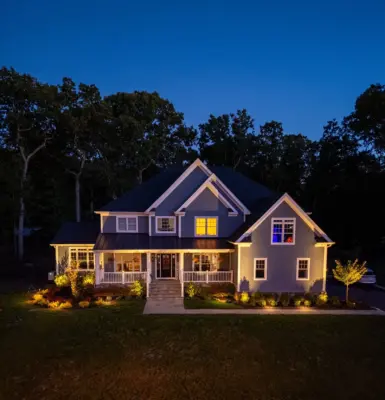 A complete outdoor transformation on a wooded lot featuring a multi-level paver patio, L-shaped outdoor kitchen, pool patio with coping, paver steps with integrated LED lighting, retaining walls, foundation plantings, and a full landscape lighting package.