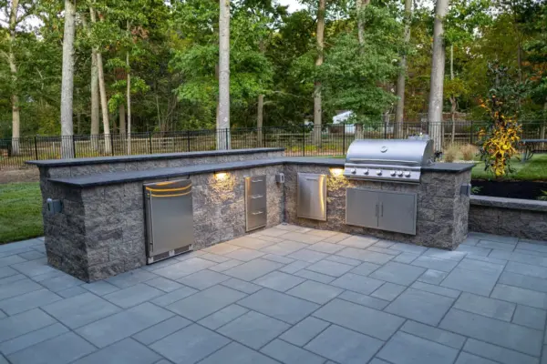 A complete outdoor transformation on a wooded lot featuring a multi-level paver patio, L-shaped outdoor kitchen, pool patio with coping, paver steps with integrated LED lighting, retaining walls, foundation plantings, and a full landscape lighting package.