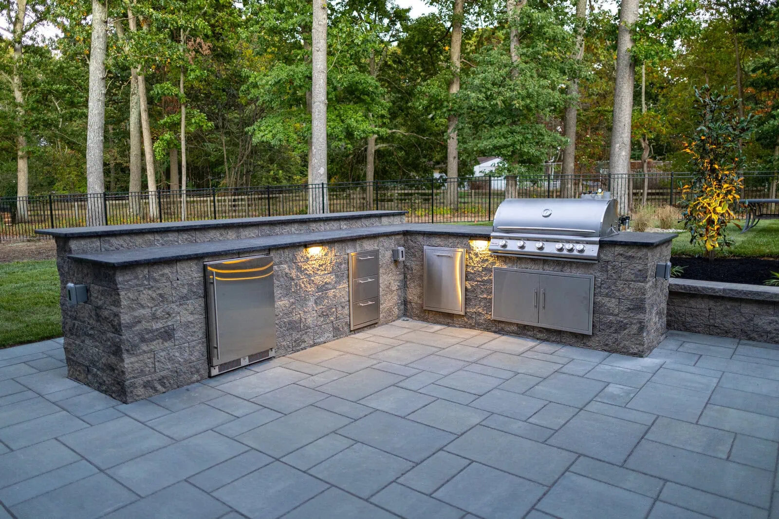 L-shaped outdoor kitchen with stone veneer, built-in stainless steel grill, refrigerator, storage drawers, and under-counter lighting on paver patio in Freehold NJ