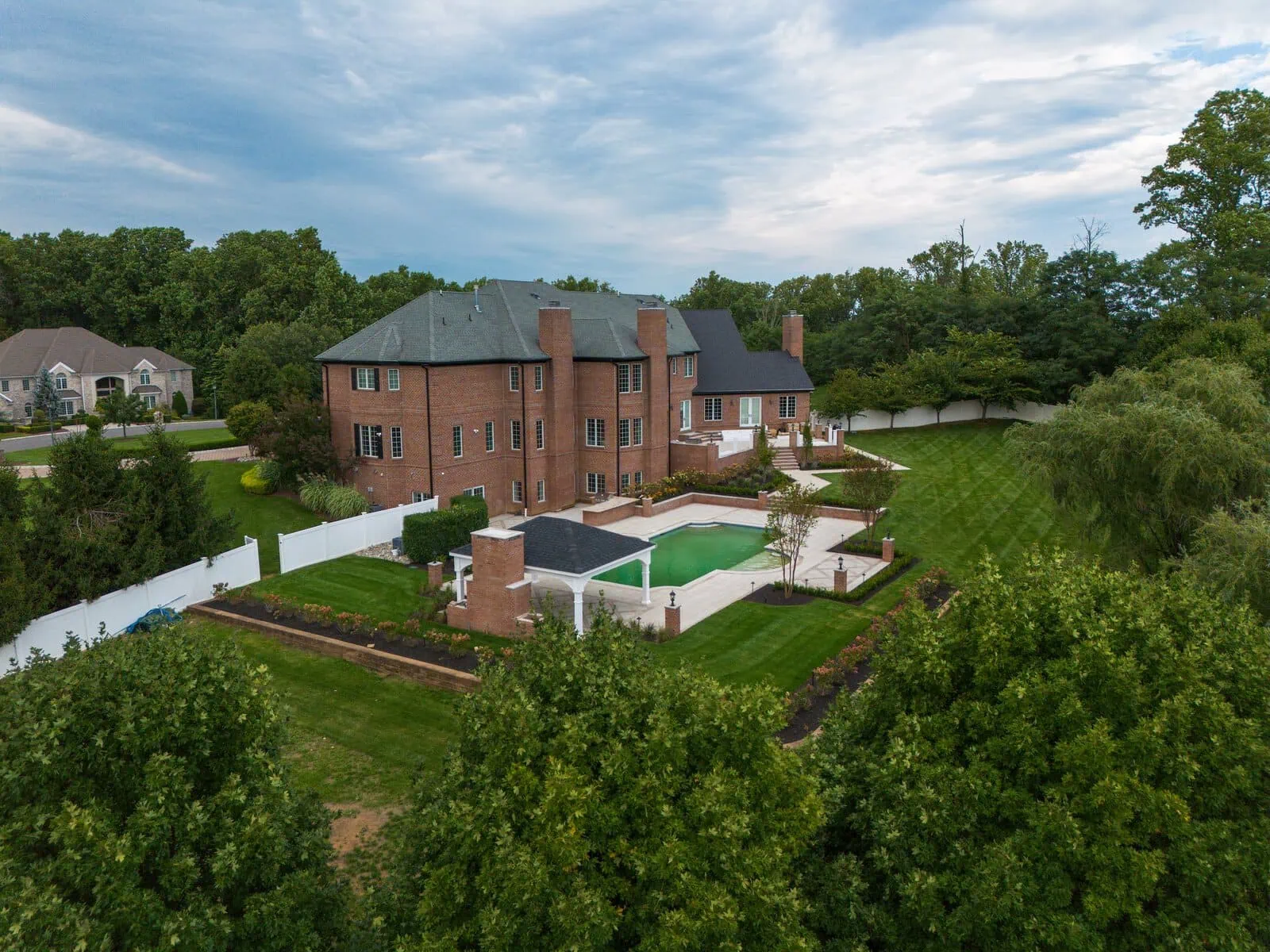 Wide aerial of a Marlboro, NJ brick estate home with pool patio, pavilion, privacy fence, and manicured lawn