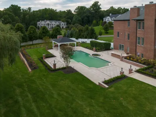 A two-level estate backyard featuring a large-format paver pool patio with decorative inlay, brick retaining walls and pillars with integrated lighting, a covered pavilion with outdoor kitchen, a side-yard paver walkway, and layered landscape plantings.