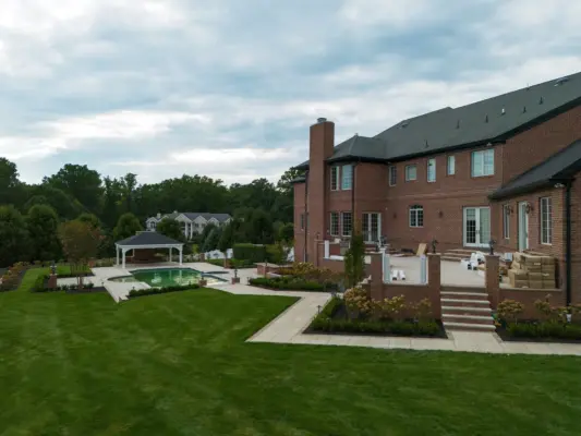 A two-level estate backyard featuring a large-format paver pool patio with decorative inlay, brick retaining walls and pillars with integrated lighting, a covered pavilion with outdoor kitchen, a side-yard paver walkway, and layered landscape plantings.