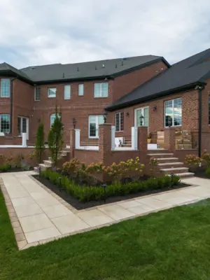 A two-level estate backyard featuring a large-format paver pool patio with decorative inlay, brick retaining walls and pillars with integrated lighting, a covered pavilion with outdoor kitchen, a side-yard paver walkway, and layered landscape plantings.