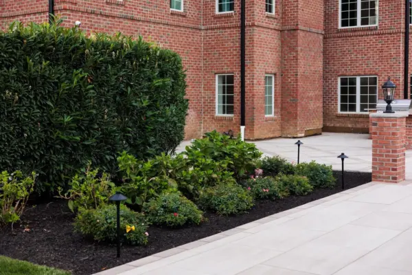 A two-level estate backyard featuring a large-format paver pool patio with decorative inlay, brick retaining walls and pillars with integrated lighting, a covered pavilion with outdoor kitchen, a side-yard paver walkway, and layered landscape plantings.