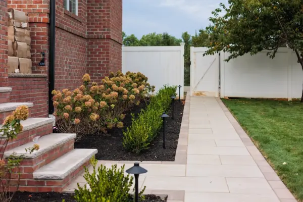 A two-level estate backyard featuring a large-format paver pool patio with decorative inlay, brick retaining walls and pillars with integrated lighting, a covered pavilion with outdoor kitchen, a side-yard paver walkway, and layered landscape plantings.