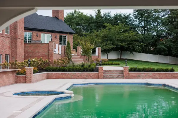 A two-level estate backyard featuring a large-format paver pool patio with decorative inlay, brick retaining walls and pillars with integrated lighting, a covered pavilion with outdoor kitchen, a side-yard paver walkway, and layered landscape plantings.