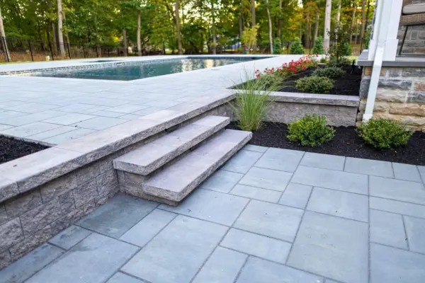 A complete outdoor transformation on a wooded lot featuring a multi-level paver patio, L-shaped outdoor kitchen, pool patio with coping, paver steps with integrated LED lighting, retaining walls, foundation plantings, and a full landscape lighting package.