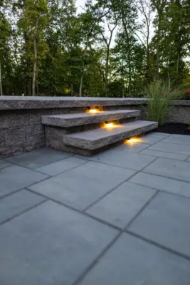A complete outdoor transformation on a wooded lot featuring a multi-level paver patio, L-shaped outdoor kitchen, pool patio with coping, paver steps with integrated LED lighting, retaining walls, foundation plantings, and a full landscape lighting package.