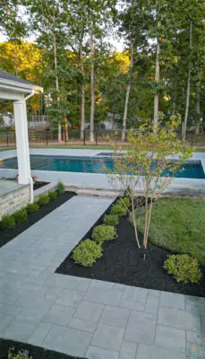 A complete outdoor transformation on a wooded lot featuring a multi-level paver patio, L-shaped outdoor kitchen, pool patio with coping, paver steps with integrated LED lighting, retaining walls, foundation plantings, and a full landscape lighting package.